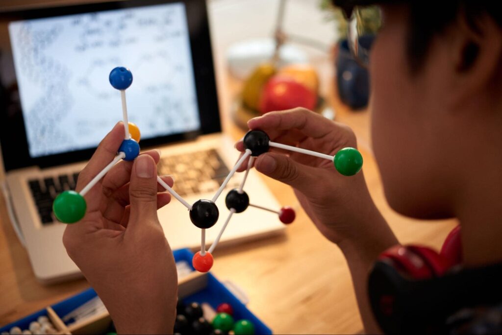 A boy is holding a model of a molecular structure in front of an open Macbook. 