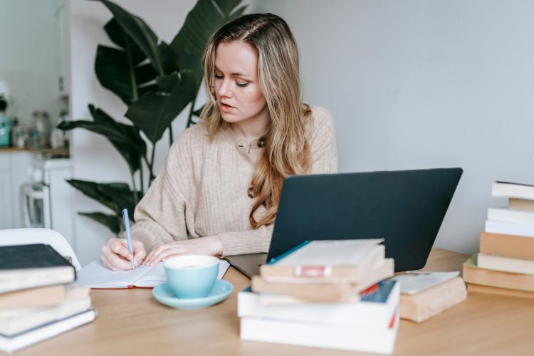 A woman writing in a notebook with an open laptop in front of her and several stacks of books around her

https://www.pexels.com/photo/pensive-businesswoman-writing-information-in-notebook-near-laptop-and-coffee-7034453/