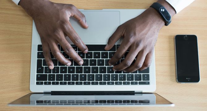 A man typing on a laptop next to his phone, both of which are propped up on a beige desk.