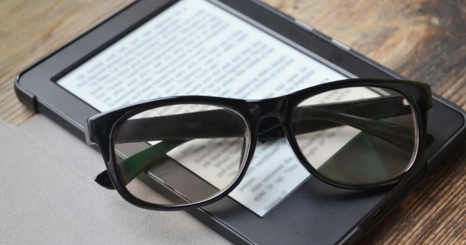 A pair of glasses on top of an e-reader tablet. Both items sit on top of a wooden table.