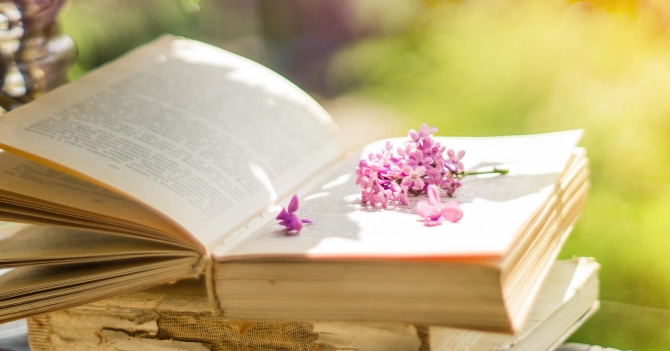 An open book with pink flowers on the page. It's set on top of a few worn books against a blurry nature background.