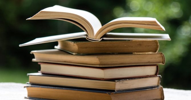 A stack of books on top of one another on a white table outdoors. The topmost book is open.