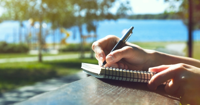 A person writing in a notebook on a table outdoors against a scenic lakeside backdrop.