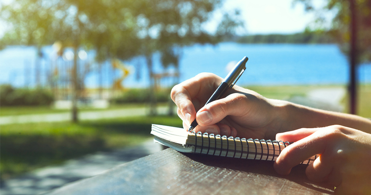 A woman sits on a bench by the lake, focused on writing in her notebook, surrounded by serene water and nature.