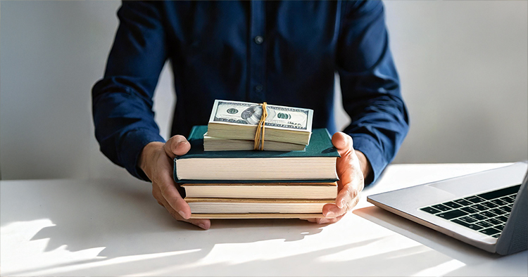 A man sitting at a desk holding a stack of books with two bill straps on top of them, asking how much does it cost to publish a book.