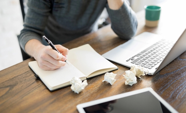 A man holding a pen over an open notebook next to a laptop, tablet, and several crumpled balls of paper.