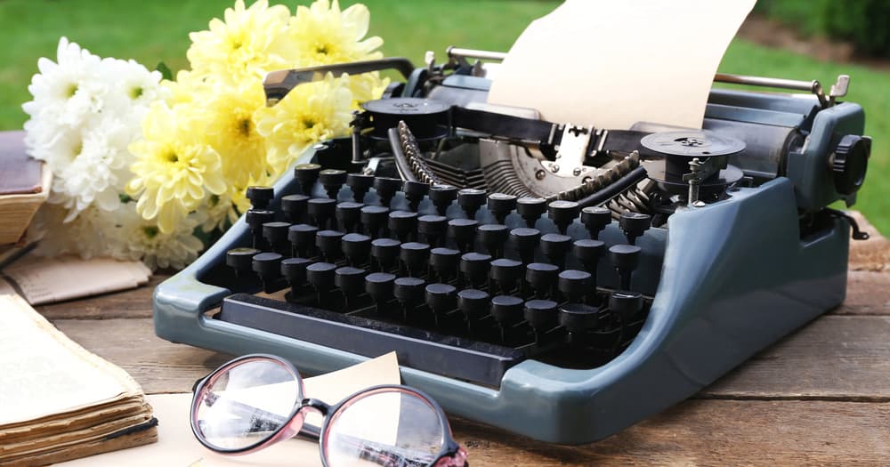 A typewriter on top of a wooden table outdoors, surrounded by flowers and books that tell you how to plot a novel.