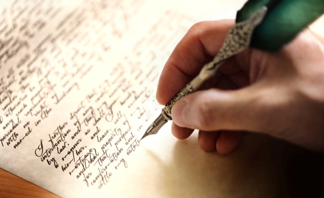 A person holding a fancy quill over a piece of paper with text written in calligraphy handwriting.